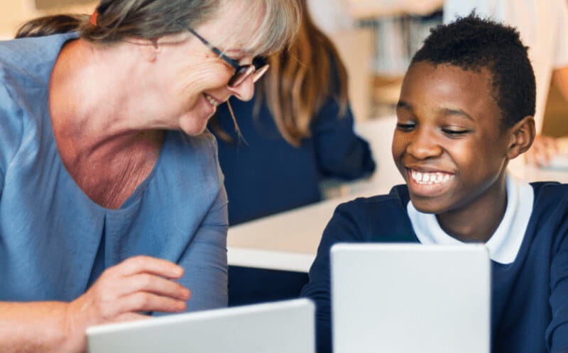 Teacher engaging with a student using a laptop in a classroom setting, exemplifying interactive learning.